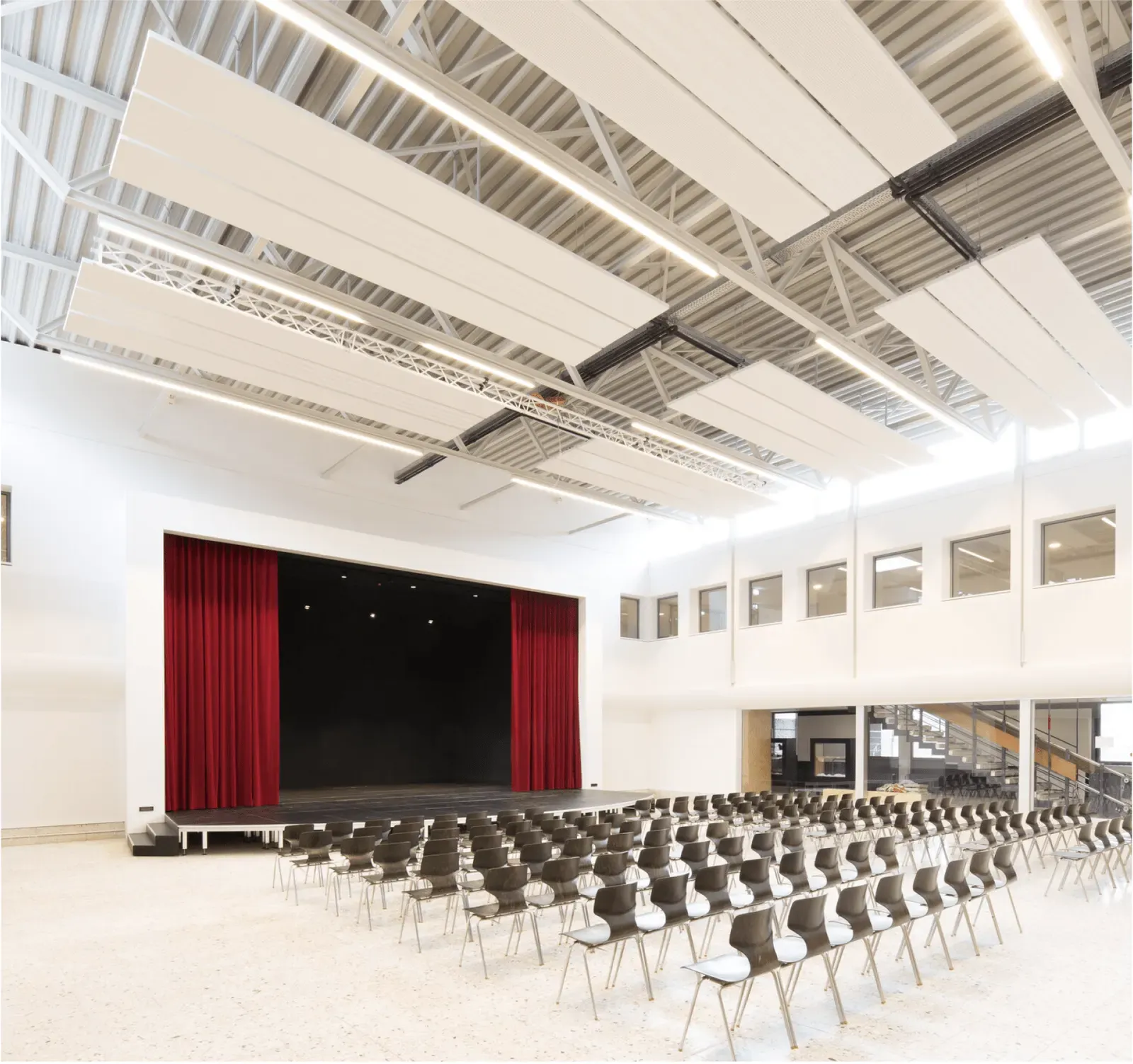 Aula mit Bühne, rotem Vorhang und Akustiksegeln unter der Decke